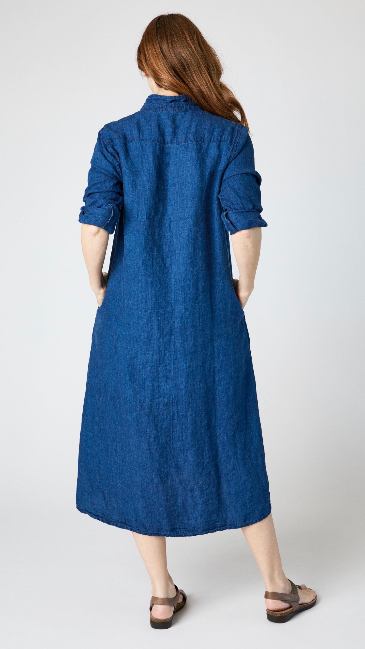 Rearview of model wearing indigo twill shirtdress with pointed collar and full length button-up panel, and brown leather sandals.