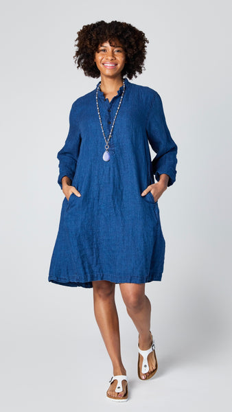 Model wearing indigo linen twill long sleeve tunic with standing collar, button-up placket and godet panels, and white leather sandals.