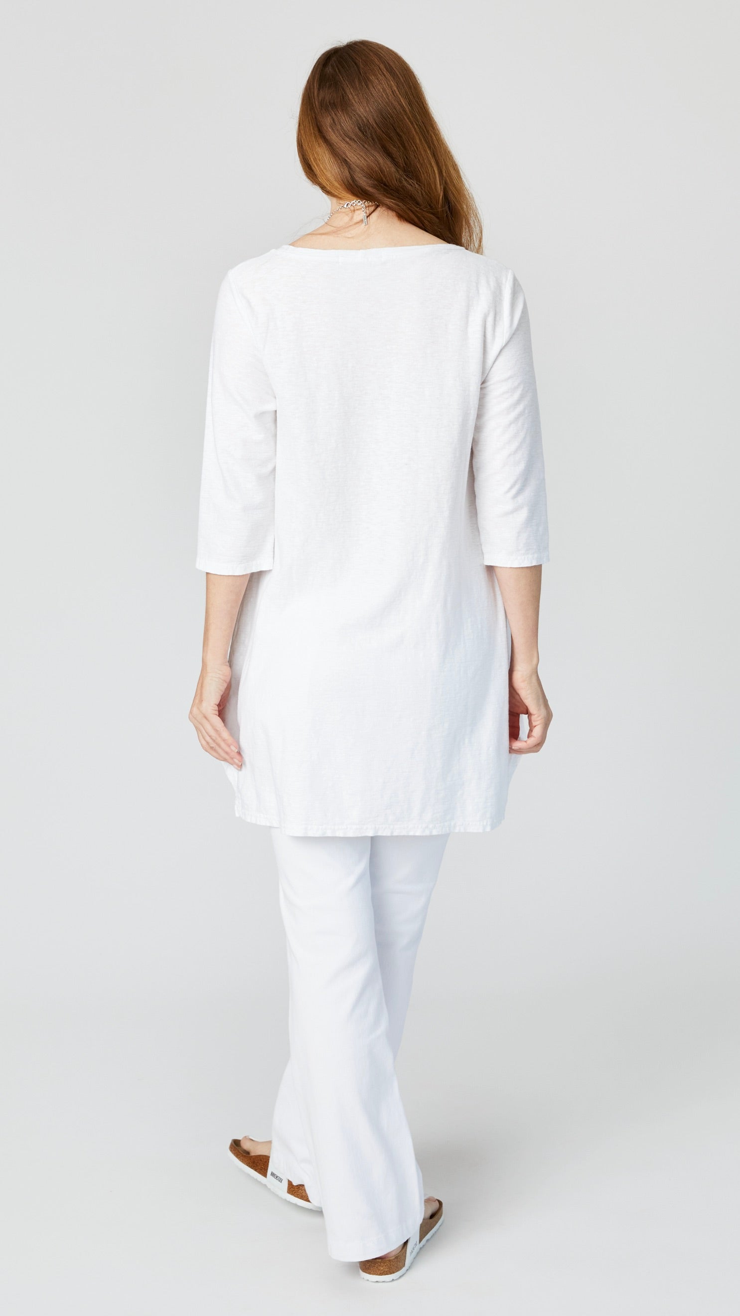 Rearview of model wearing white 3/4 sleeve high-low tunic top, white bootcut jeans, and white sandals.