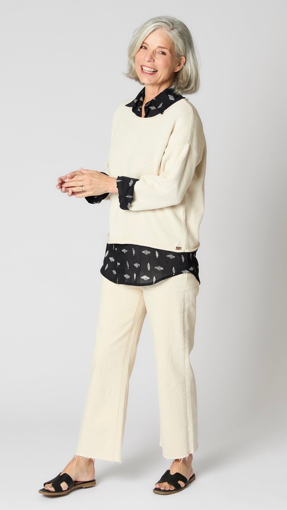 Model wearing cream boxy cotton sweater with drop shoulder and slightly cropped hemline, layered over black button-up shirt, and cropped ecru wideleg jeans.