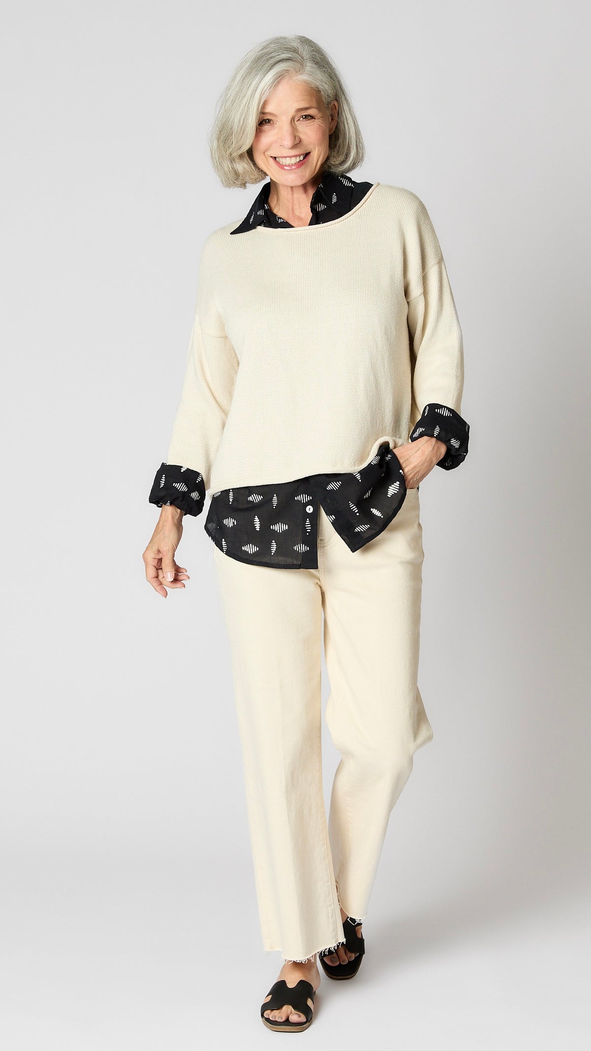 Model wearing cream boxy cotton sweater with drop shoulder and slightly cropped hemline, layered over black button-up shirt, and cropped ecru wideleg jeans.