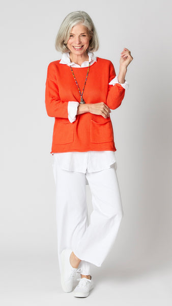 Model wearing sunset coral cotton sweater with boatneck and patch pockets, white linen button-up top, and white wide-leg cropped jeans.