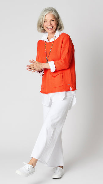 Model wearing sunset coral cotton sweater with boatneck and patch pockets, white linen button-up top, and white wide-leg cropped jeans.