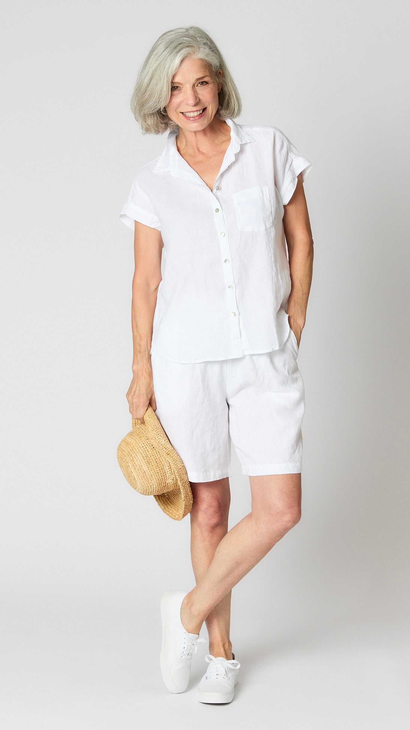 Model wearing white linen capsleeve camp shirt with mother of pearl buttons, and knee length white linen shorts.