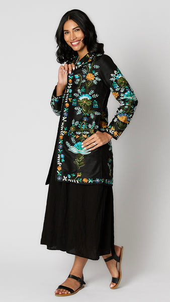 Model wearing black silk duster embroidered with bright blue, yellow and green tones over black rayon romper and black leather sandals.