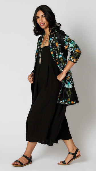 Model wearing black silk duster embroidered with bright blue, yellow and green tones over black rayon romper and black leather sandals.