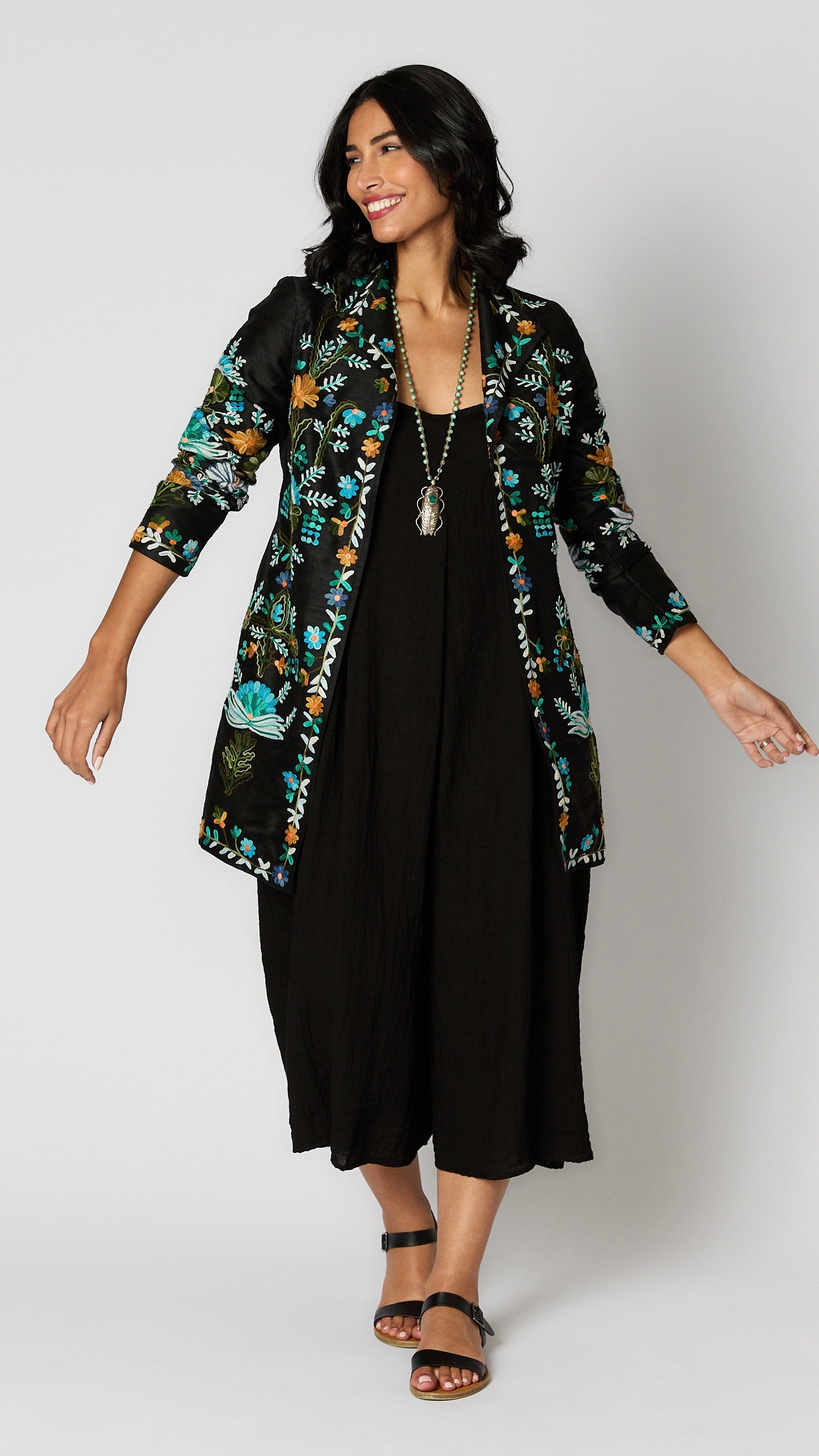 Model wearing black silk duster embroidered with bright blue, yellow and green tones over black rayon romper and black leather sandals.