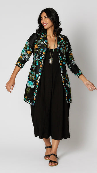 Model wearing black silk duster embroidered with bright blue, yellow and green tones over black rayon romper and black leather sandals.