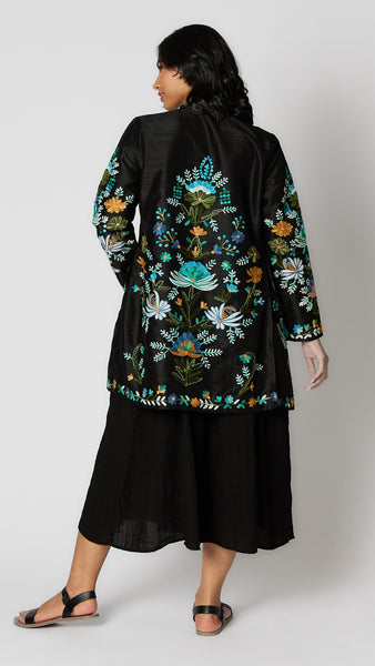 Model wearing black silk duster embroidered with bright blue, yellow and green tones over black rayon romper and black leather sandals.