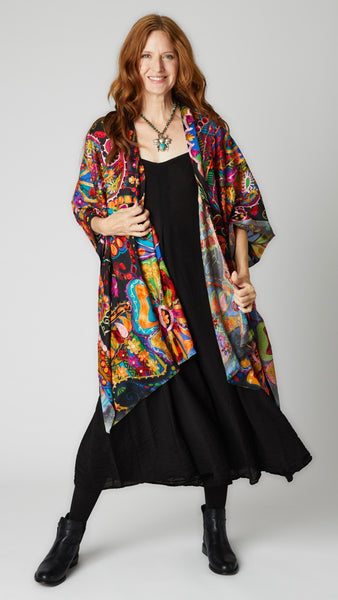 Model wearing multicolor/rainbow ruana with black background over black easy romper and black leather boots.