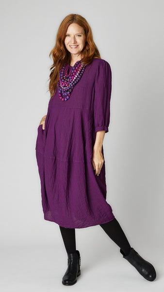 Model wearing mulberry mid-calf length rayon dress with black leggings and black leather boots. Dress has scoop neck, 3/4 sleeves, side seam pockets and slight tulip silhouette.