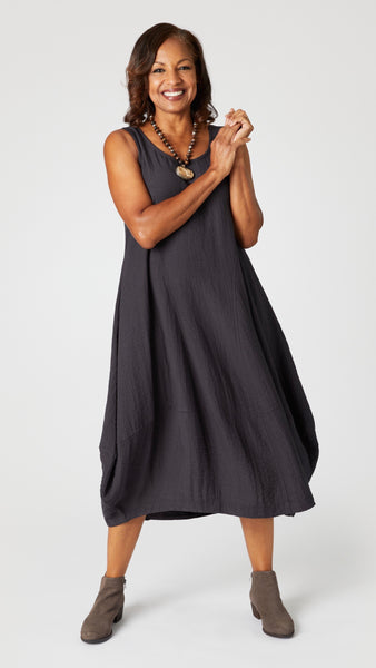 Model wearing "black pearl" sleeveless rayon dress with tulip hemline and round neck, beaded necklace with bone pendant, and gray suede boots.