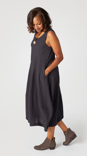 Model wearing "black pearl" sleeveless rayon dress with tulip hemline and round neck, beaded necklace with bone pendant, and gray suede boots.