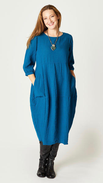 Model wearing deep sea mid-calf length rayon dress with black leggings and black leather boots. Dress has scoop neck, 3/4 sleeves, side seam pockets and slight tulip silhouette.
