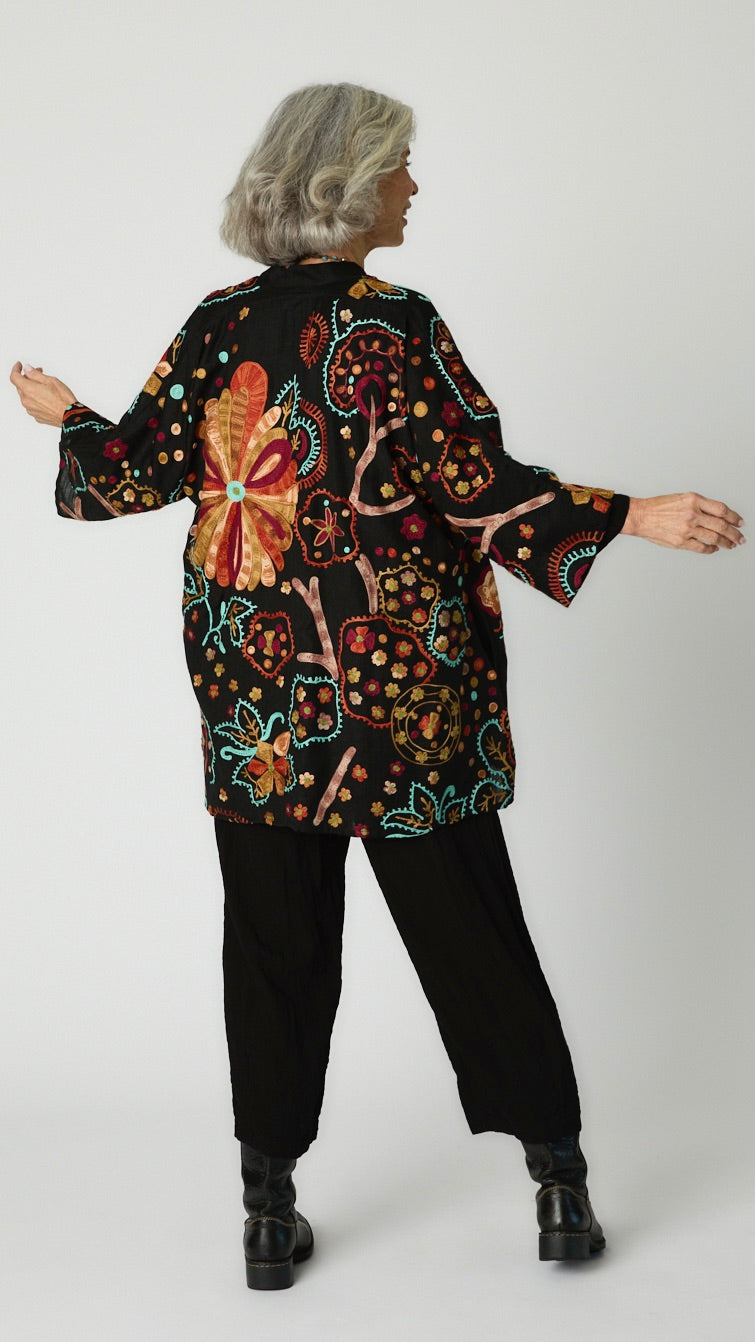 Rearview of model in black cotton jacket with floral embroidery shows rich orange, gold, red tones and vibrant turquoise floral patterns.