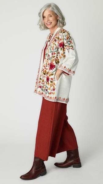 Model wearing white silk kimono style jacket with red, green, yellow floral embroidery, cognac cropped top and cognac cropped dart pants in rayon, carved bone rabbit pendant necklace, and brown leather boots.