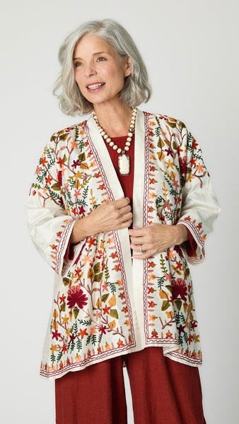 Model wearing white silk kimono style jacket with red, green, yellow floral embroidery, cognac cropped top and cognac cropped dart pants in rayon, carved bone rabbit pendant necklace.