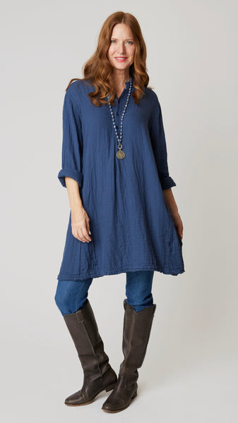 Model wearing indigo linen twill long sleeve tunic with standing collar, button-up placket and godet panels, midwash denim leggings, and leather boots.