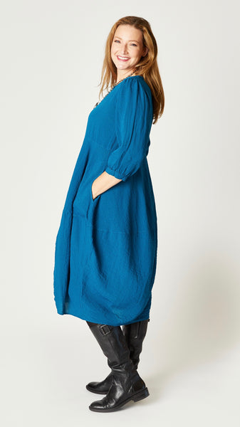 Model wearing deep sea mid-calf length rayon dress with black leggings and black leather boots. Dress has scoop neck, 3/4 sleeves, side seam pockets and slight tulip silhouette.