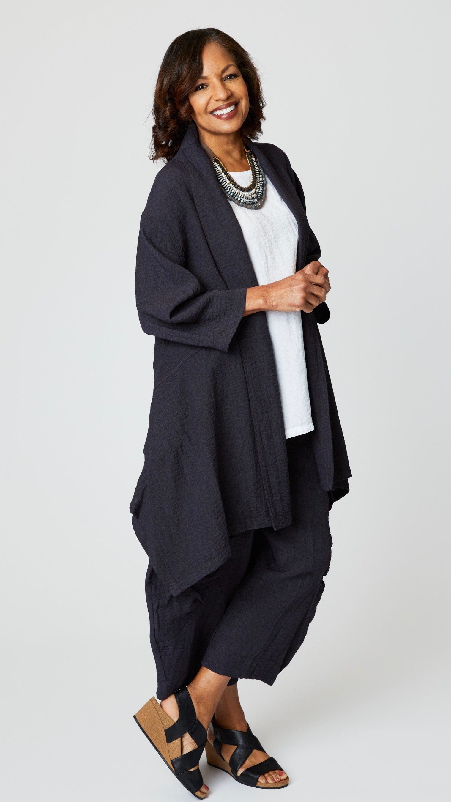 Model wearing "black pearl" rayon jacket with shawl collar, 3/4 sleeves, and tunic length hemline over white long tank, cropped "black pearl" double pocket rayon pant, and black leather sandals.