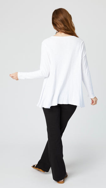 Rearview of model wearing white jersey long sleeve top with hip length flare and round neckline, black bootcut jeans, and black leather sandals.