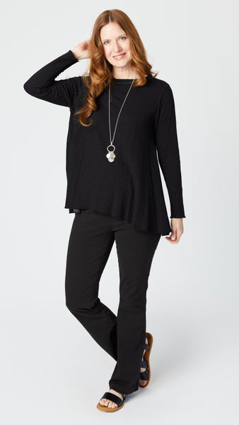 Rearview of model wearing black jersey long sleeve top with hip length flare and round neckline, black bootcut jeans, and black leather sandals.