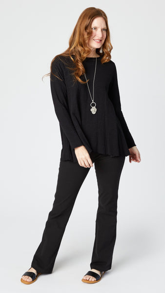 Rearview of model wearing black jersey long sleeve top with hip length flare and round neckline, black bootcut jeans, and black leather sandals.