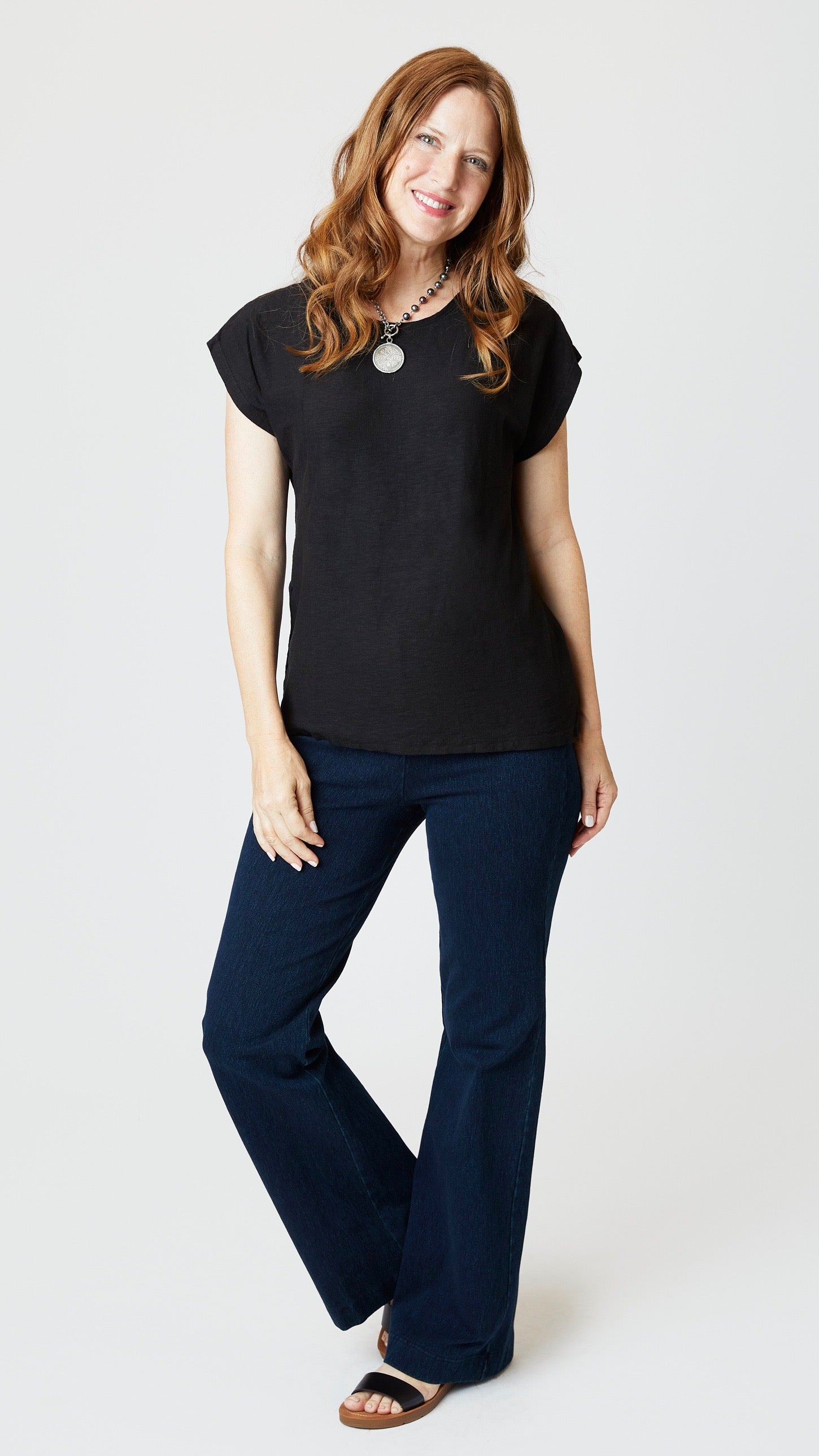 Model wearing black cotton-linen jersey top with cropped sleeves, boatneck, and boxy silhouette with indigo bootcut jeans, and sandals.