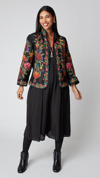 Model wearing shorter black silk jacket embroidered in shades of dusty pink, purple, orange, and green over a black rayon romper, black tights, and black leather ankle boots.