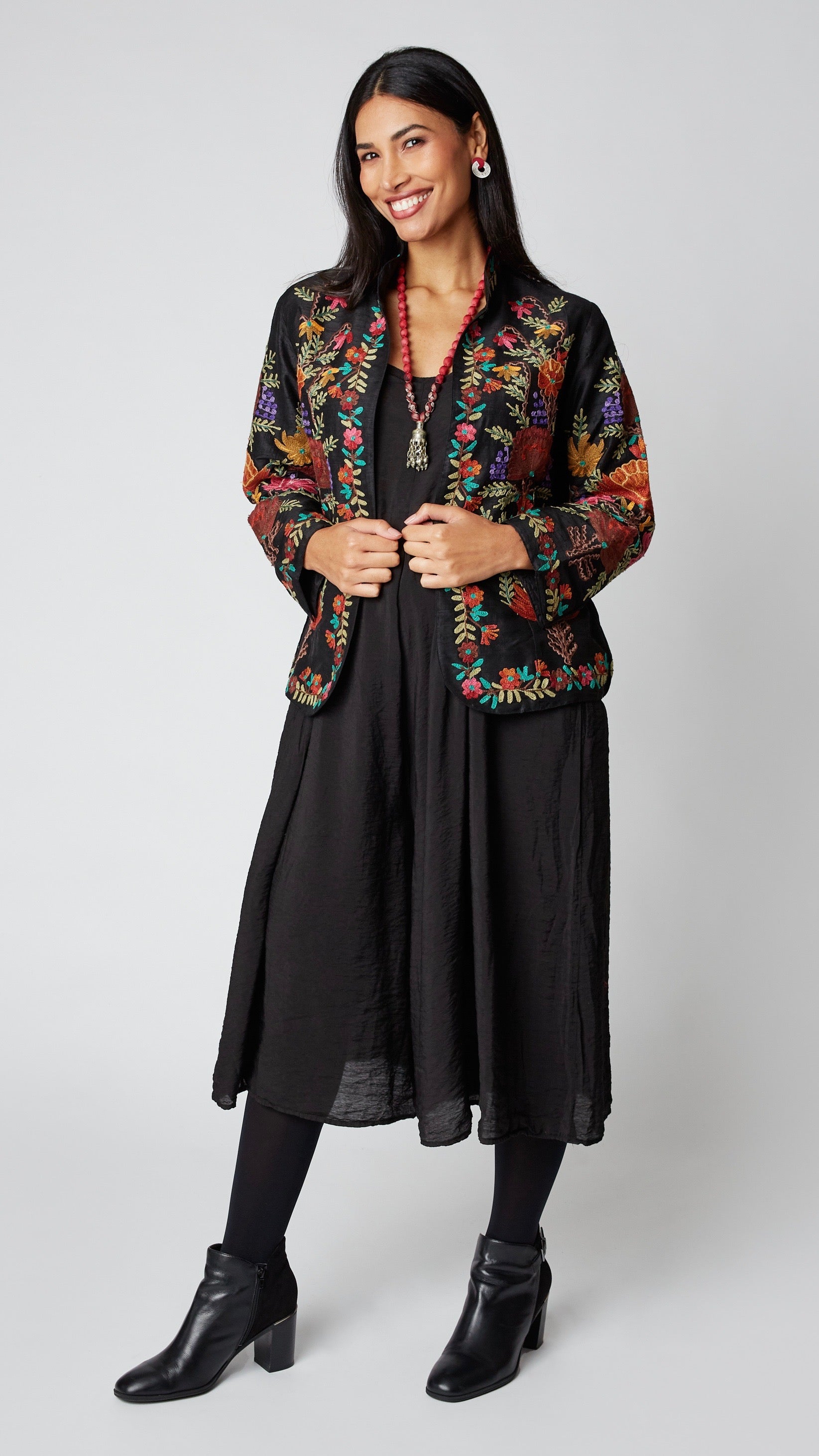 Model wearing shorter black silk jacket embroidered in shades of dusty pink, purple, orange, and green over a black rayon romper, black tights, and black leather ankle boots.