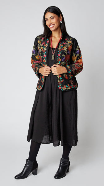 Model wearing shorter black silk jacket embroidered in shades of dusty pink, purple, orange, and green over a black rayon romper, black tights, and black leather ankle boots.