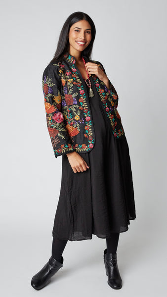 Model wearing shorter black silk jacket embroidered in shades of dusty pink, purple, orange, and green over a black rayon romper, black tights, and black leather ankle boots.