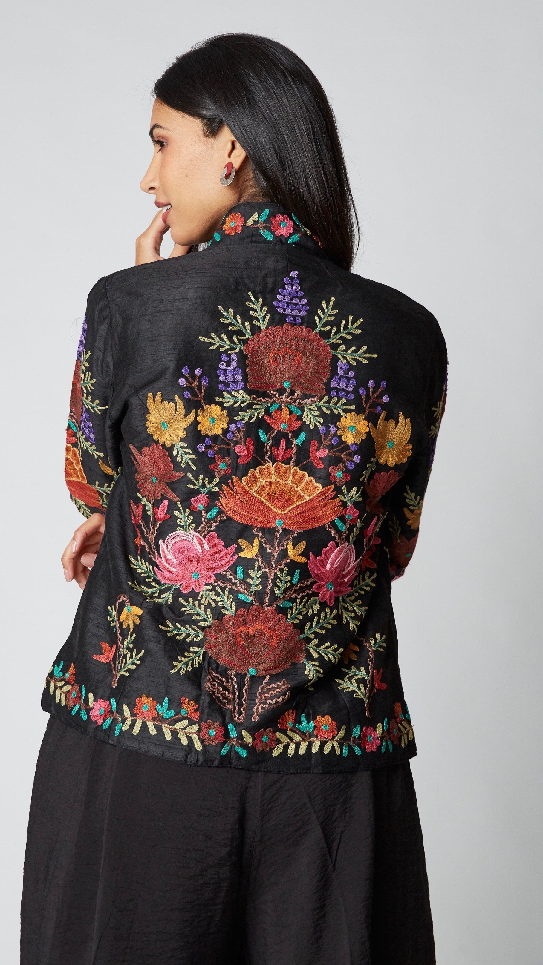 Model wearing shorter black silk jacket embroidered in shades of dusty pink, purple, orange, and green over a black rayon romper, black tights, and black leather ankle boots.