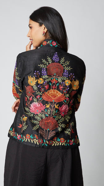 Model wearing shorter black silk jacket embroidered in shades of dusty pink, purple, orange, and green over a black rayon romper, black tights, and black leather ankle boots.