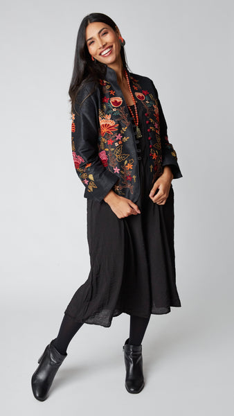 Model wearing shorter black silk jacket embroidered in shades of dusty pink, red, orange, and brown over a black rayon romper, black tights, and black leather ankle boots.