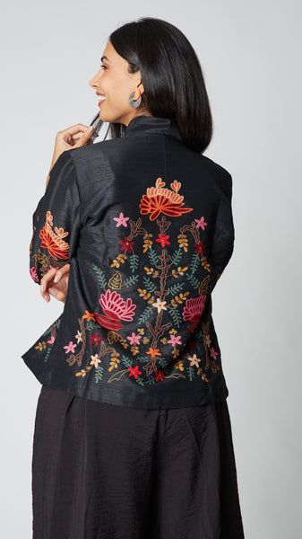 Model wearing shorter black silk jacket embroidered in shades of dusty pink, red, orange, and brown over a black rayon romper, black tights, and black leather ankle boots.