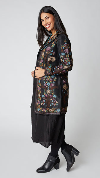Model wearing black silk duster embroidered in dusty blue, purple, and brown tones over black rayon romper and black suede boots.