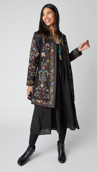 Model wearing black silk duster embroidered in dusty blue, purple, and brown tones over black rayon romper and black suede boots.
