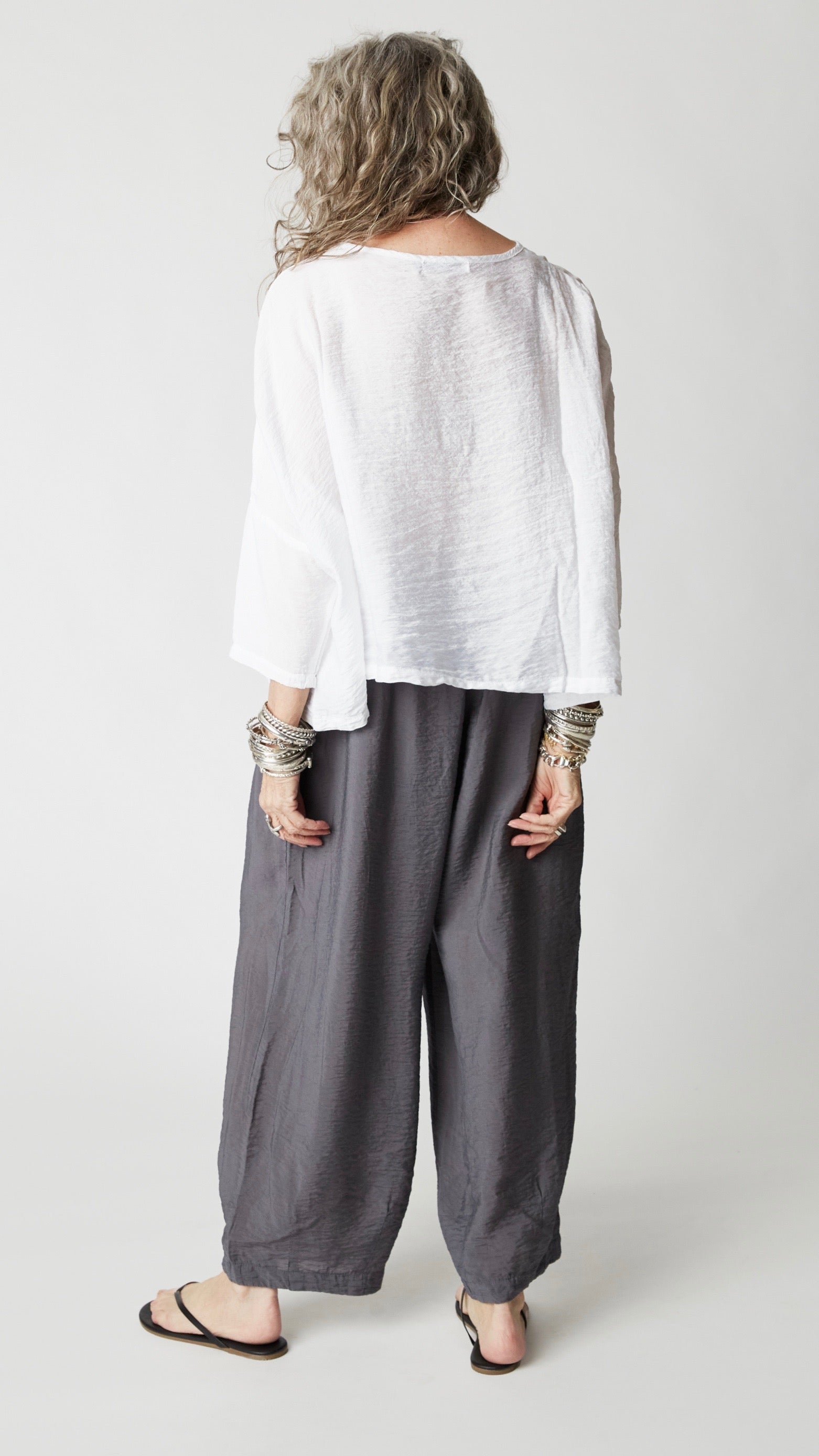 Model wearing cropped white rayon top with high round neckline, dropped shoulders and ¾ sleeves, gray cropped dart pants, silver bangles, and black flip-flops.