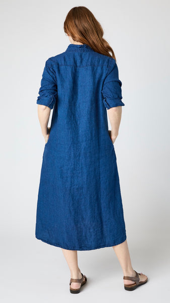 Rearview of model wearing indigo twill shirtdress with pointed collar and full length button-up panel, and brown leather sandals.