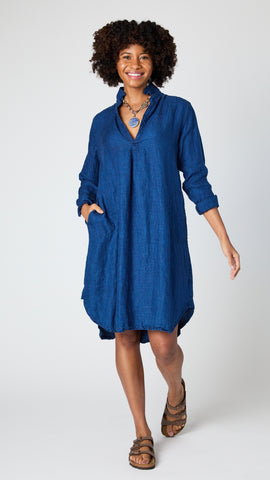 Model wearing indigo twill long sleeve dress with deep V-neck, collar, and shirttail hem, necklace with lapis pendant, and brown leather sandals.