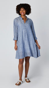 Model wearing blue chambray linen long sleeve tunic with standing collar, button-up placket and godet panels, and white leather sandals.