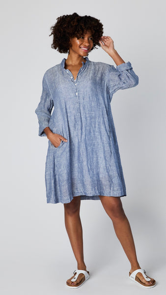 Model wearing blue chambray linen long sleeve tunic with standing collar, button-up placket and godet panels, and white leather sandals.