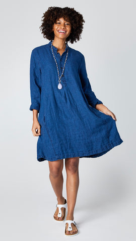 Model wearing indigo linen twill long sleeve tunic with standing collar, button-up placket and godet panels, and white leather sandals.
