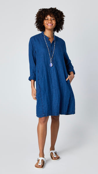 Model wearing indigo linen twill long sleeve tunic with standing collar, button-up placket and godet panels, and white leather sandals.