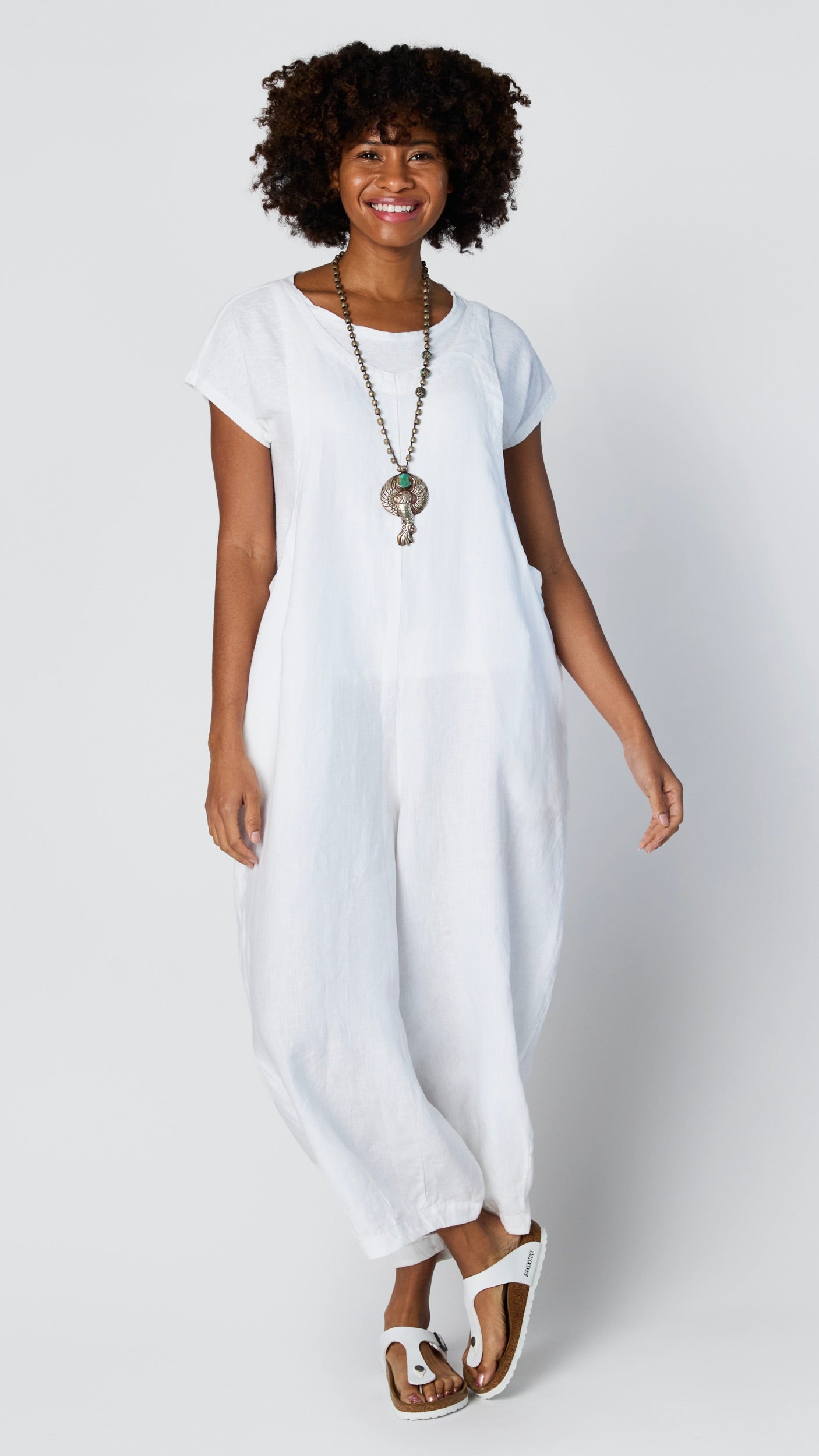 Model wearing white linen overall jumpsuit with wide cropped dart leg, white 3/4 sleeve bias cut tee, and white sandals.