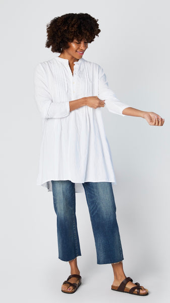 Model wearing white cotton twill tunic with front button, long sleeves, pleating at yoke, and cropped wideleg midwash jeans.