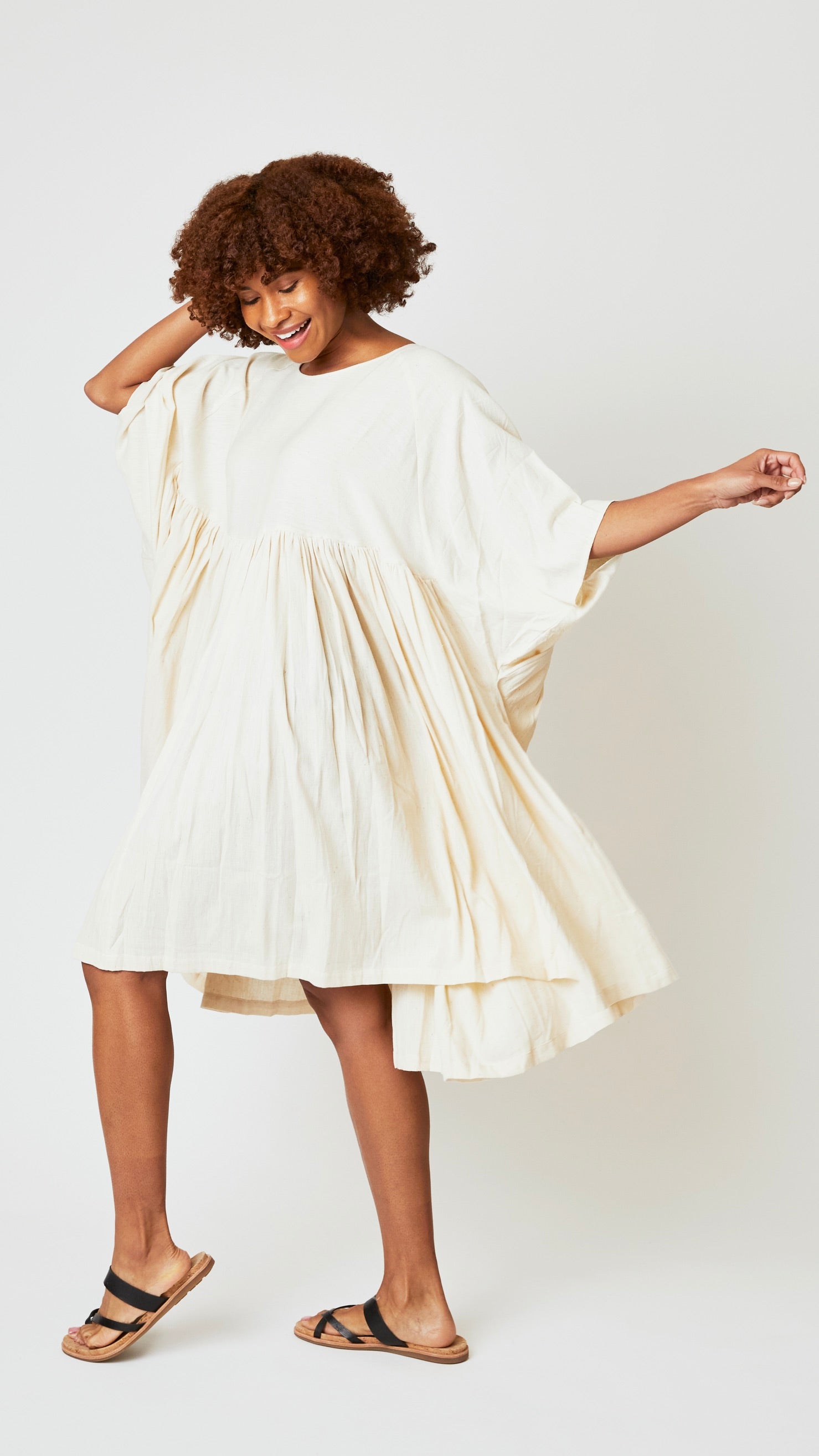 Model wearing oversized natural cotton dress with raglan sleeves and waist-gathered skirt, and black leather sandals.
