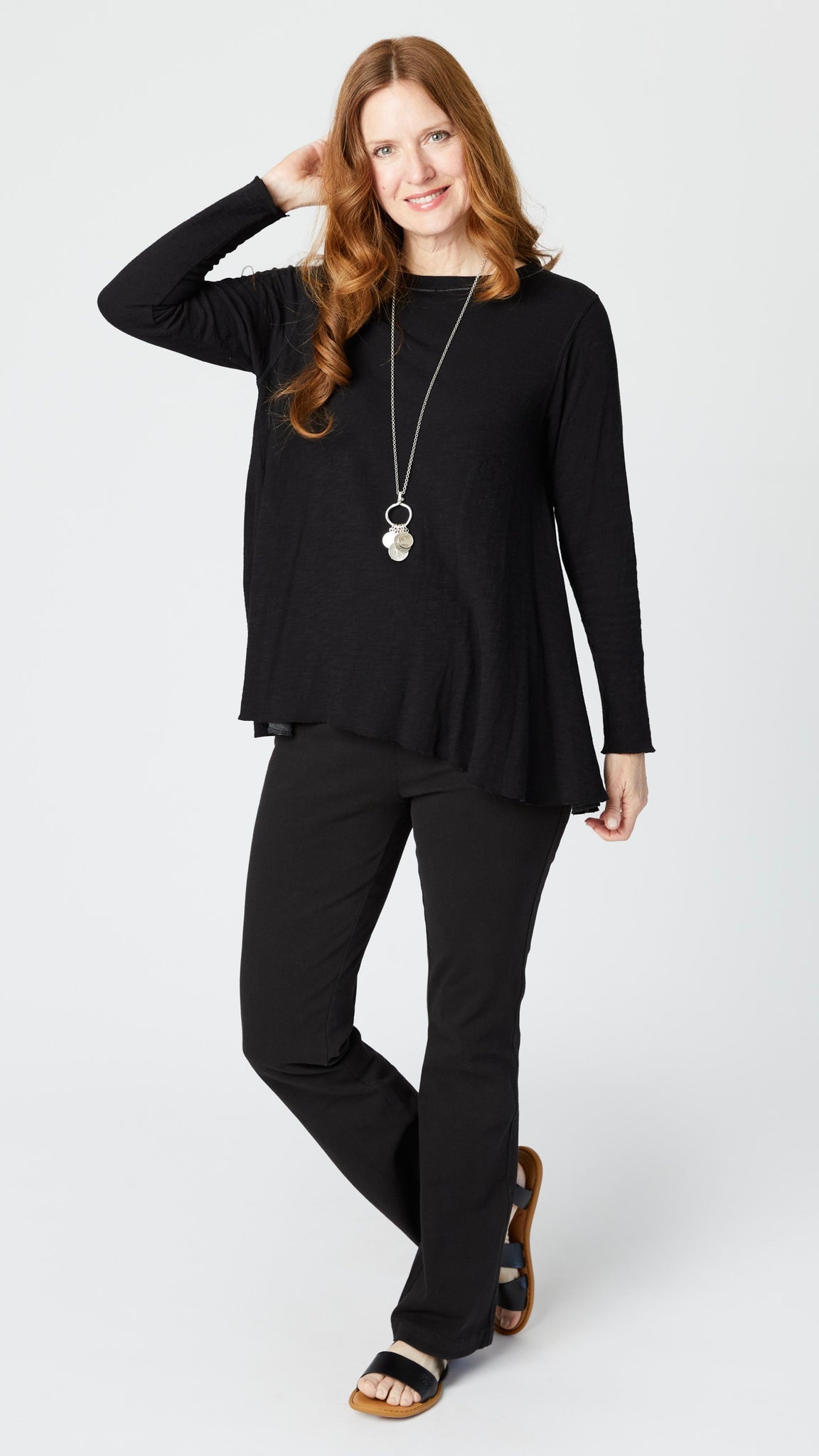 Model wearing black long sleeve swing top, black bootcut denim jeans, silver pendant necklace, and black leather sandals.