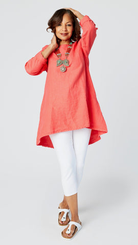 Model wearing coral red high-low linen tunic with long sleeves, white capri leggings, coral and turquoise necklace, and white sandals.